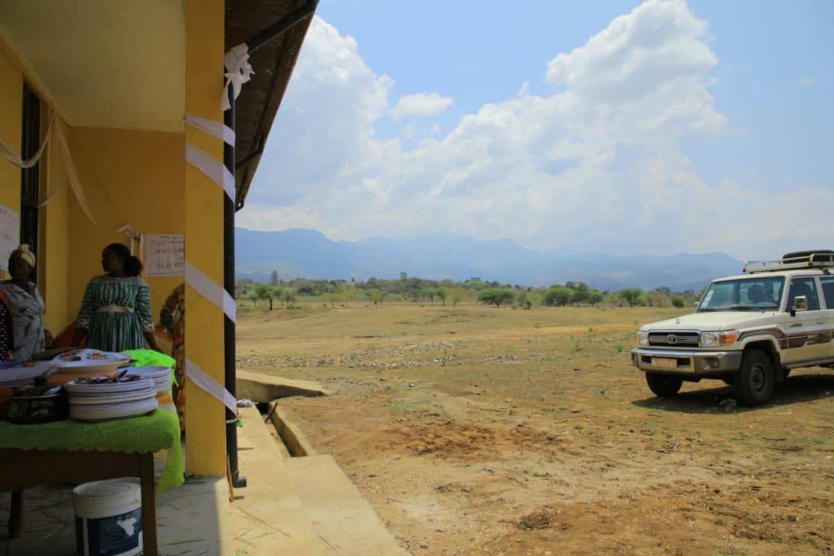 Feest in Bele Town met mensen en een voertuig in een uitgestrekt landschap.