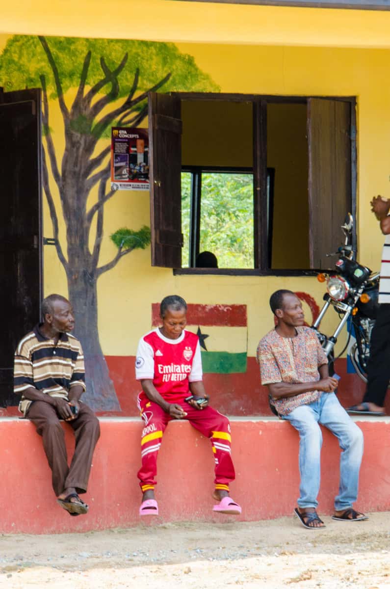 Mensen wachten buiten een lokaal met een kleurrijke muur en een boommuurkunst.